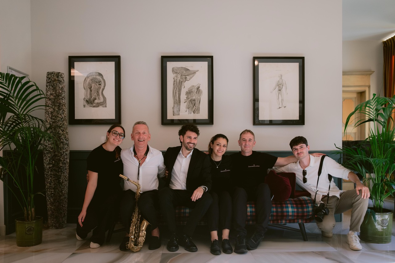 The team and partners of Extraordinary Weddings sitting together on a bench in a villa lounge, with a saxophone and a camera