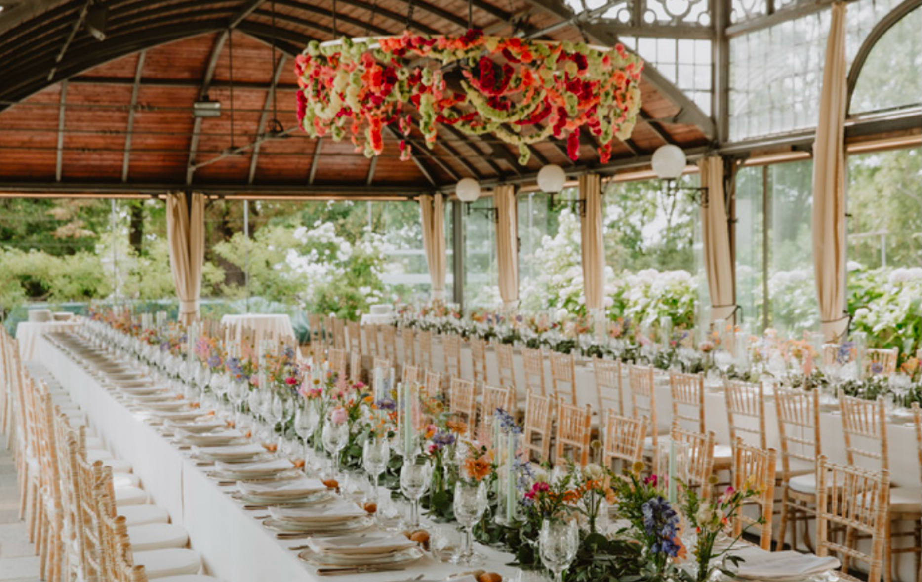 Long banquet table dressed with flowers under a glasshouse roof and a hanging floral installation